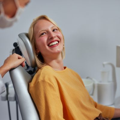 Female dentist wearing medical overalls, mask and gloves talking to young woman lying on dental chair